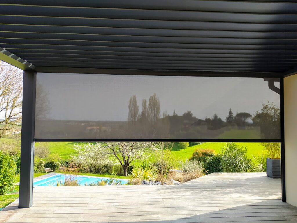 Vue sous une pergola bioclimatique moderne avec store extérieur baissé, donnant sur une terrasse bois, piscine et jardin verdoyant.