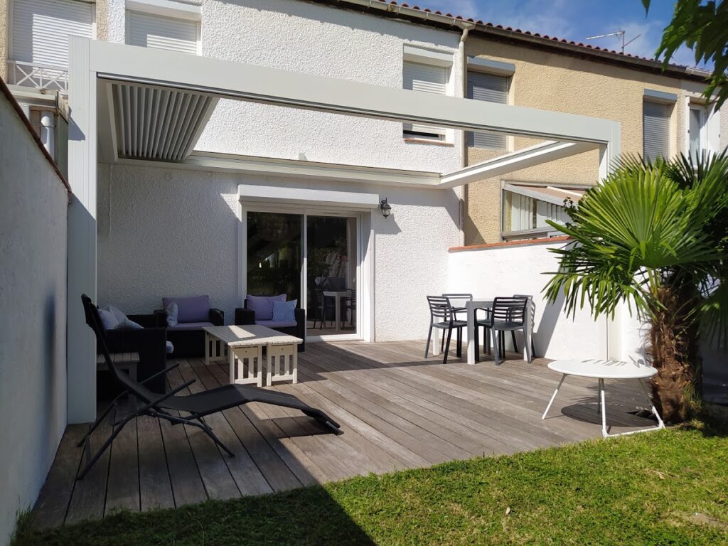 Terrasse bois avec pergola bioclimatique blanche, salon de jardin et salle à manger extérieure devant une maison.