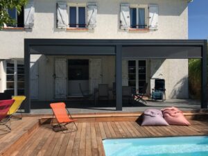 Maison blanche avec pergola grise couvrant une terrasse aménagée. Piscine avec deck bois, transats colorés et poufs.