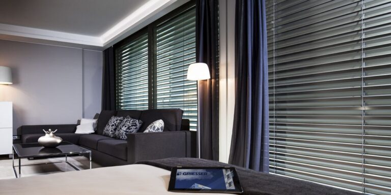 Salon confortable avec canapé, stores à lamelles et rideaux devant grande fenêtre, tablette GRIESSER sur plaid.