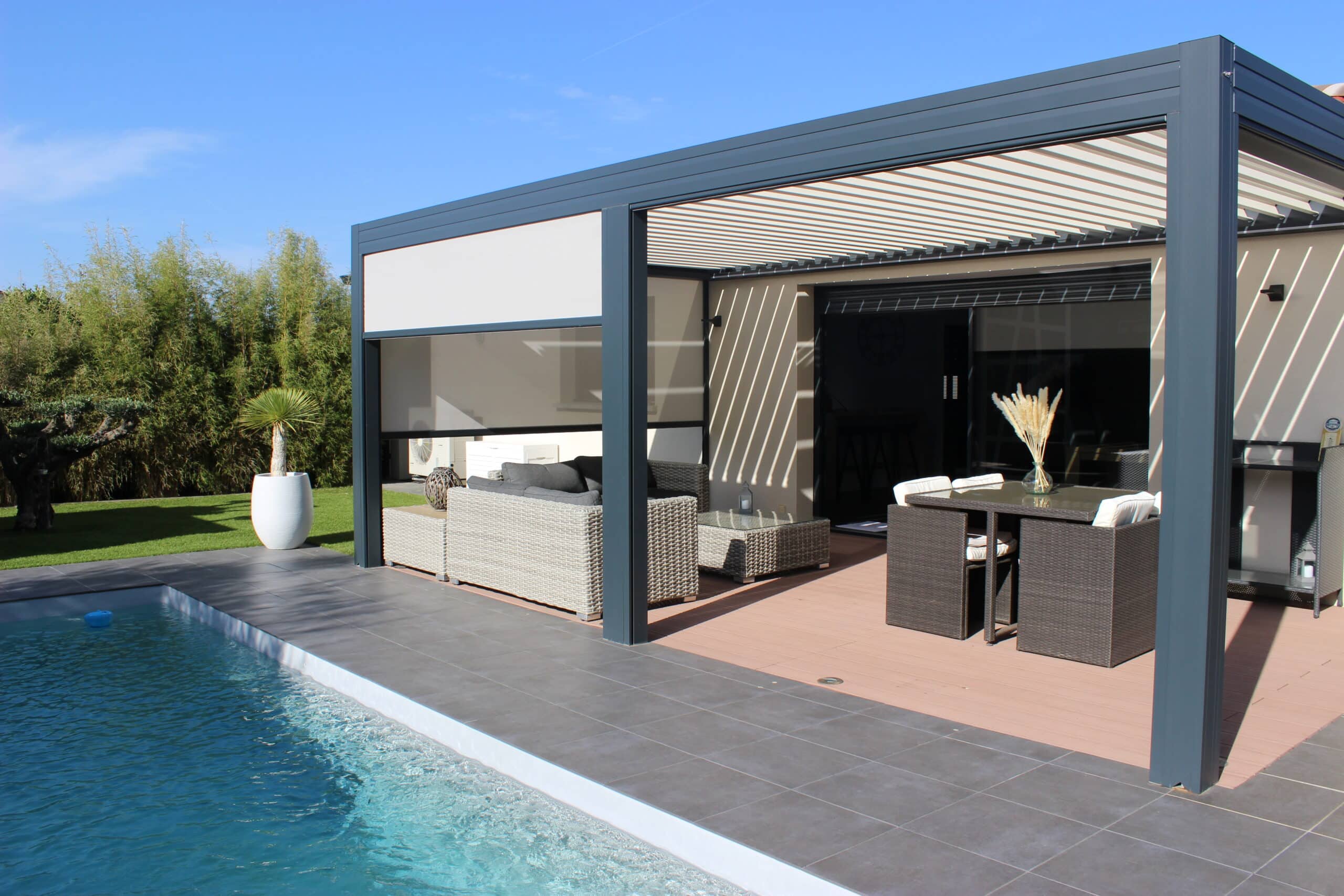 Grande pergola bioclimatique gris foncé, stores screens latéraux abaissés. Espace lounge sur terrasse bois et piscine carrelée.