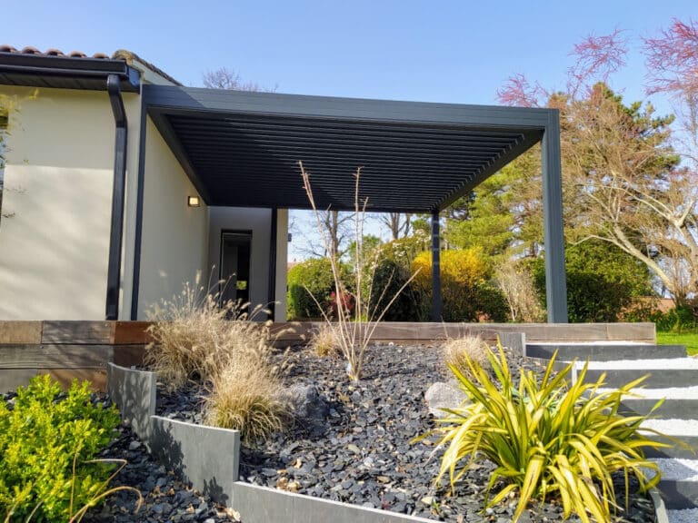 Pergola bioclimatique moderne gris anthracite devant une maison claire, avec jardin rocheux, paillis d'ardoise et plantes ornementales.
