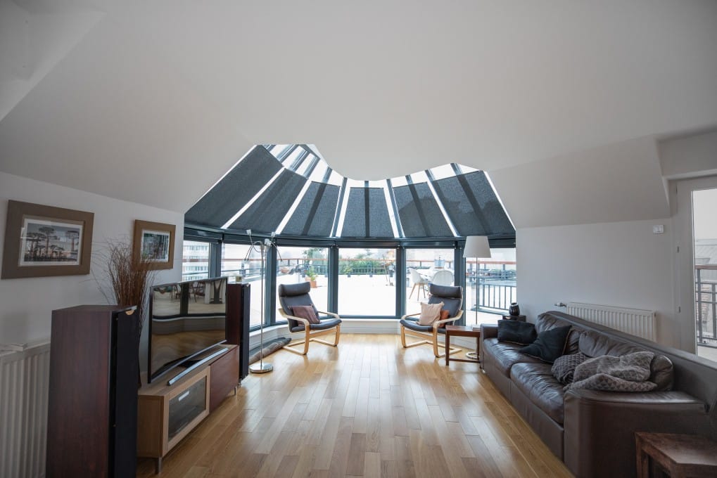 Salon lumineux avec grandes fenêtres panoramiques équipées de stores. Le mobilier inclut un canapé en cuir, deux fauteuils et une TV.