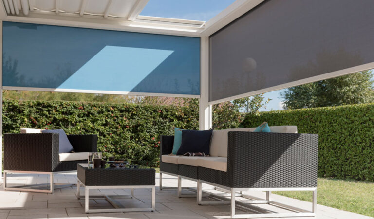 Patio sous pergola moderne avec stores latéraux bleus et gris. Salon de jardin confortable devant une haie verte dense.