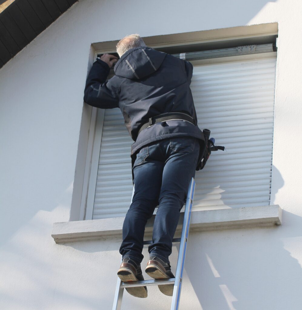 Homme sur une échelle réparant le mécanisme d'un volet roulant sur une fenêtre. Mur extérieur blanc ensoleillé.