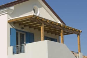 Façade blanche méditerranéenne avec pergola en bois, balcon à balustrade métal et volets bleus sous un ciel clair.