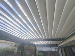 Pergola bioclimatique moderne avec lattes orientables partiellement ouvertes, dévoilant un ciel bleu parsemé de nuages. Structure sombre.