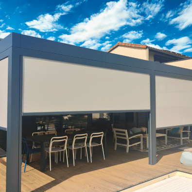 Pergola moderne grise avec stores verticaux blancs abaissés, couvrant une terrasse en bois avec salle à manger extérieure.