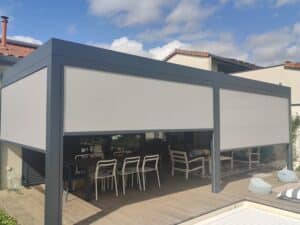 Pergola moderne avec stores latéraux gris clair abaissés. Un salon de jardin et une table à manger sur terrasse en bois, ciel bleu nuageux.