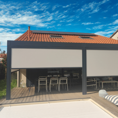 Pergola bioclimatique grise protégeant une terrasse en bois grâce à deux grands stores latéraux beiges. Salle à manger d'été en dessous.