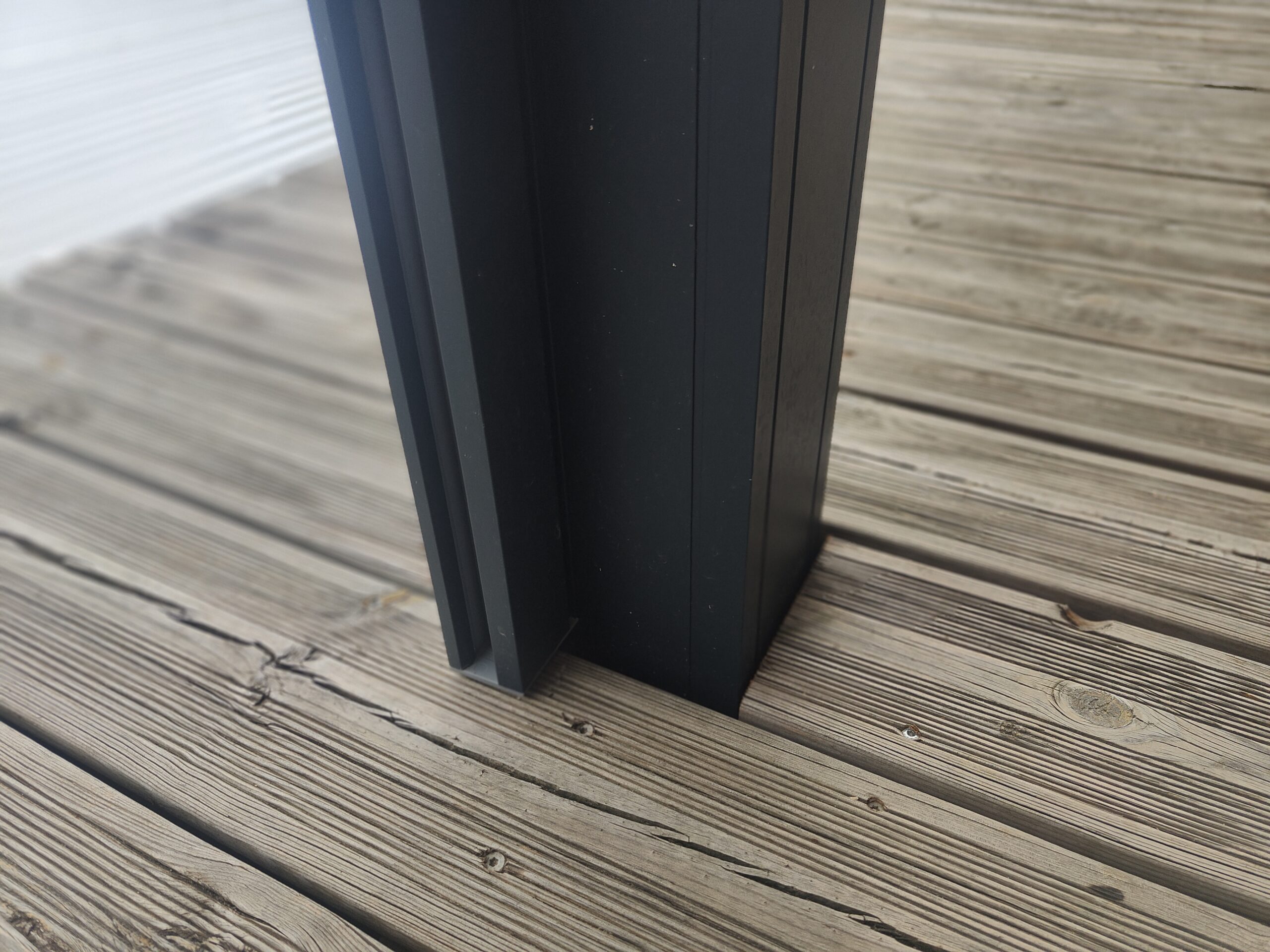 Poteau anthracite moderne sur terrasse en bois avec planches rainurées et vis apparentes. Contraste entre métal sombre et bois clair.