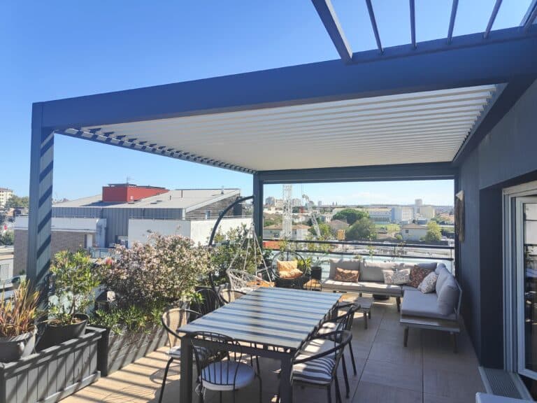Terrasse de toit moderne avec pergola bioclimatique, salon et salle à manger d'extérieur. Vue sur la ville sous ciel bleu.