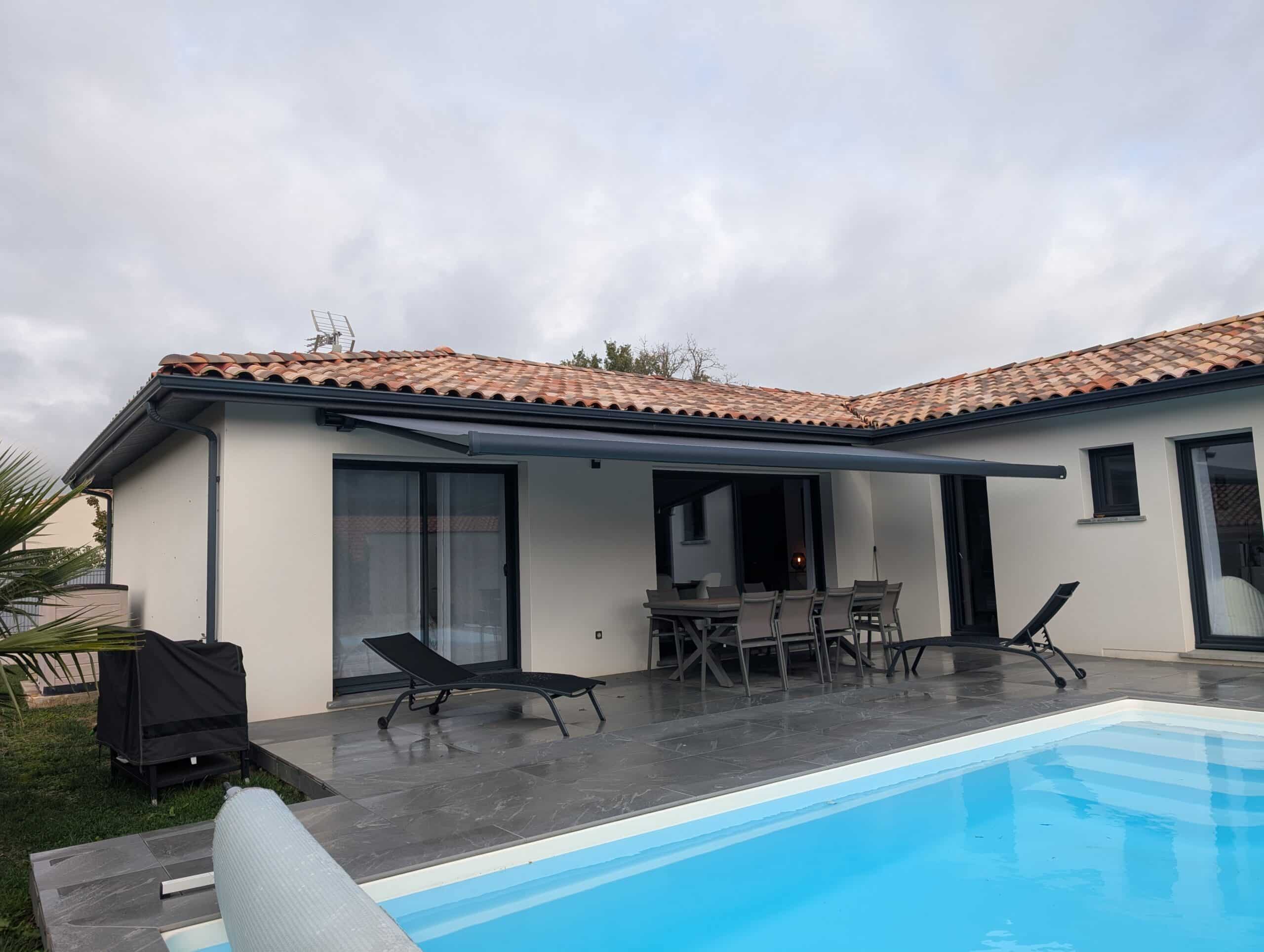 Maison moderne avec store banne déplié, terrasse carrelée sombre et piscine aux eaux bleues sous un ciel gris et nuageux.