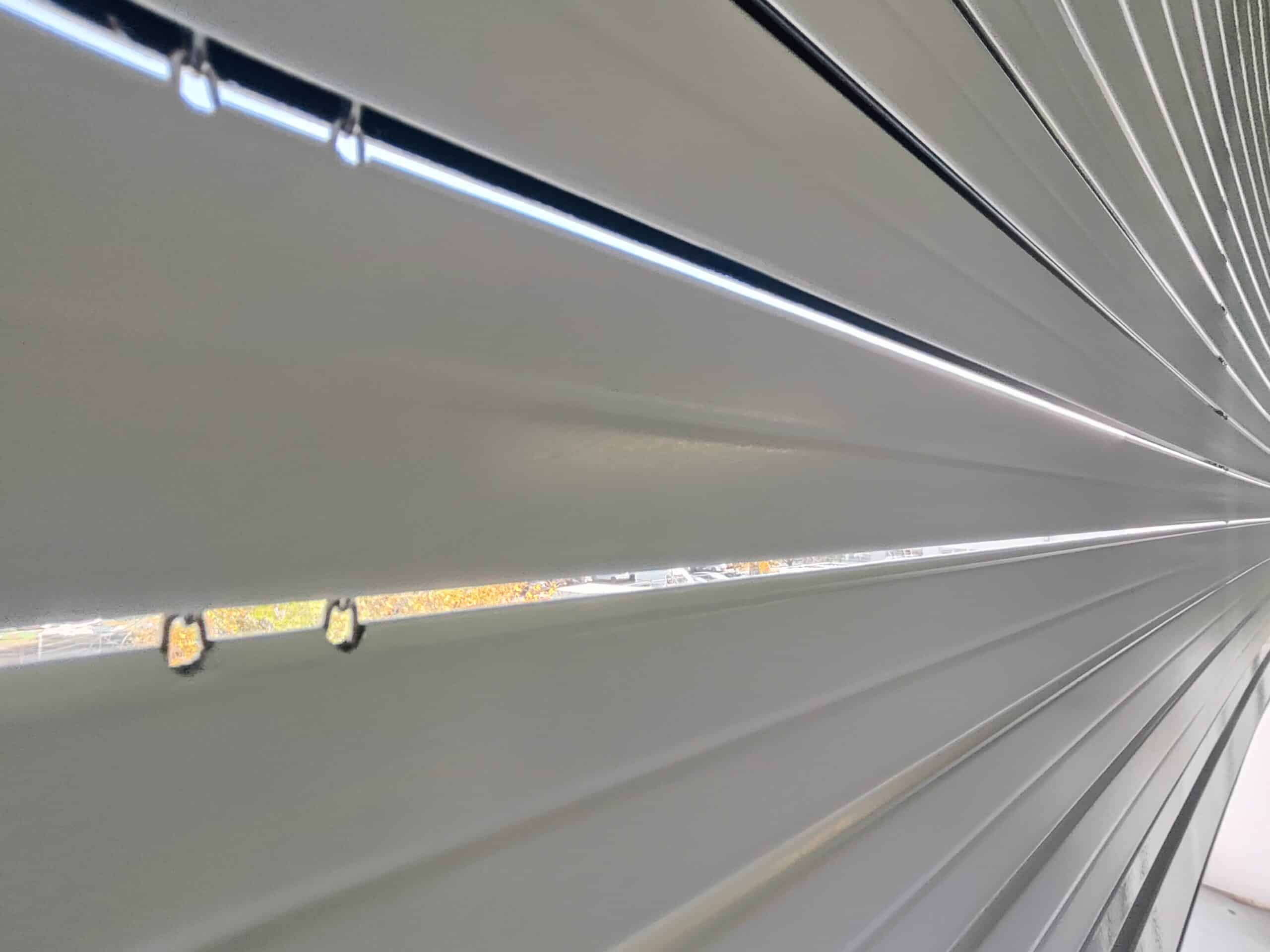 Vue rapprochée en plongée d'un volet roulant gris clair à lamelles, laissant entrevoir une ligne de lumière et un feuillage jaune à l'extérieur.