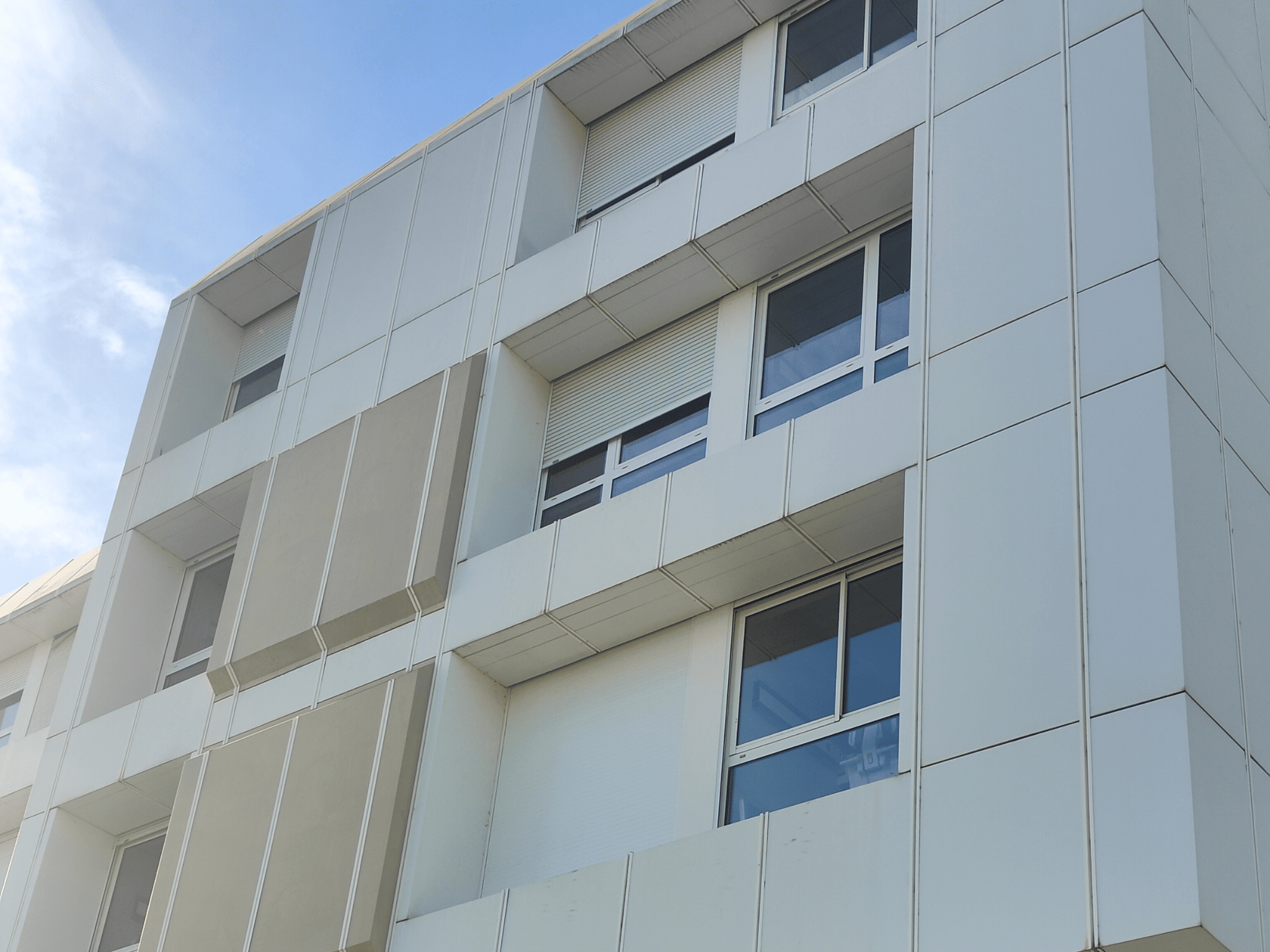 Façade moderne d'un immeuble avec des panneaux blancs et beiges, fenêtres carrées et stores roulants partiellement baissés sous un ciel bleu clair.