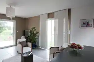 Salon moderne avec fauteuils blancs, tapis shaggy, et grandes baies vitrées occultées par des stores à panneaux blancs.