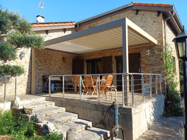 Grande terrasse de maison en pierre avec pergola bioclimatique moderne et toit à lames orientables. Meubles de jardin en bois et garde-corps en inox.