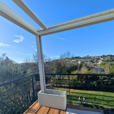 Pergola blanche sur terrasse bois, poteau intégré dans une grande jardinière. Vue ensoleillée sur la colline verdoyante et les maisons.