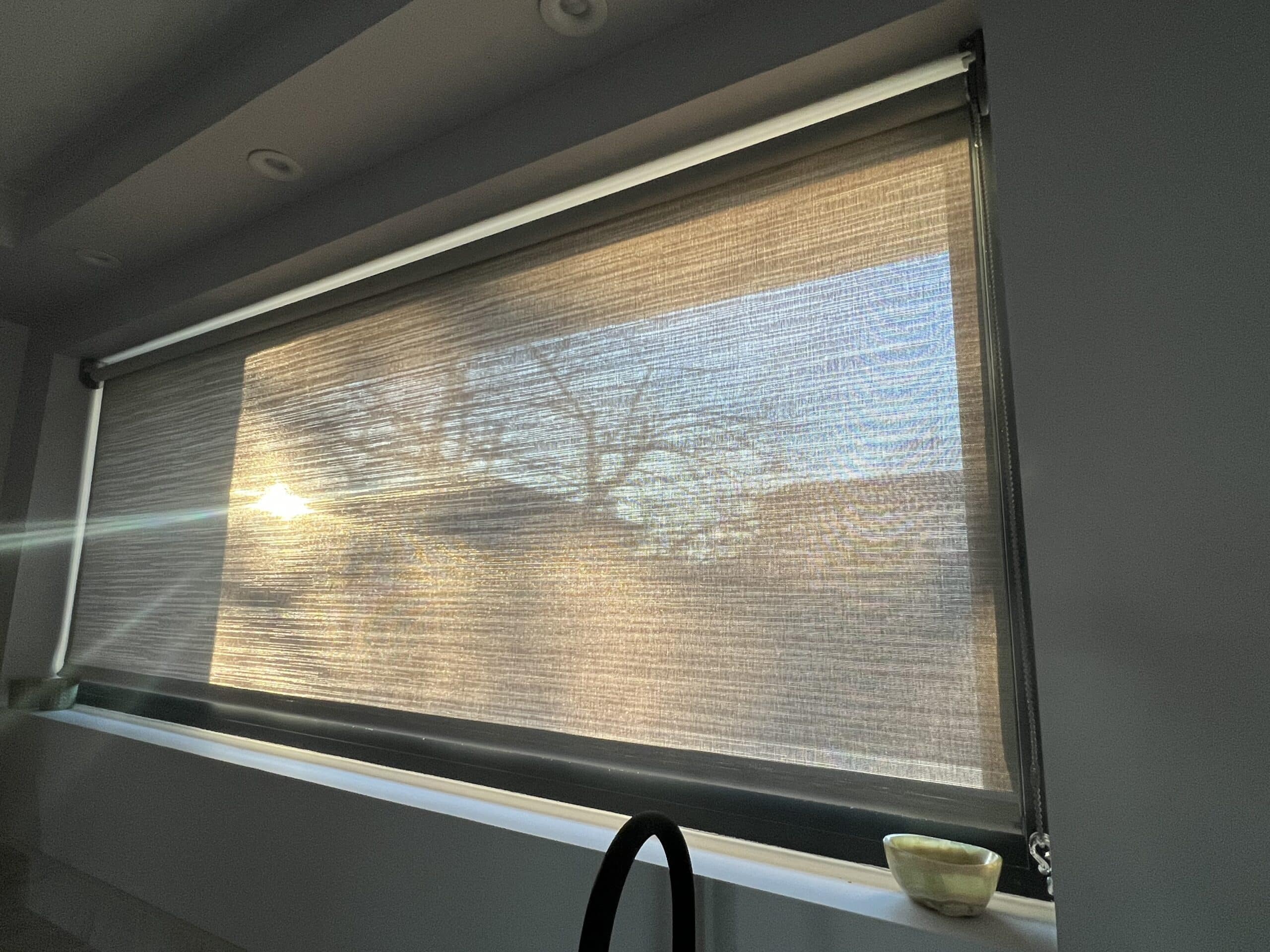 Store enrouleur tamisant la forte lumière du soleil couchant sur une large fenêtre. L'écran révèle la silhouette des branches d'arbre.