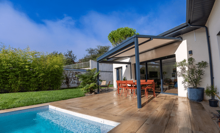 Terrasse en bois clair bordant une piscine bleue. Une pergola moderne protège une salle à manger extérieure orange dans un jardin verdoyant.