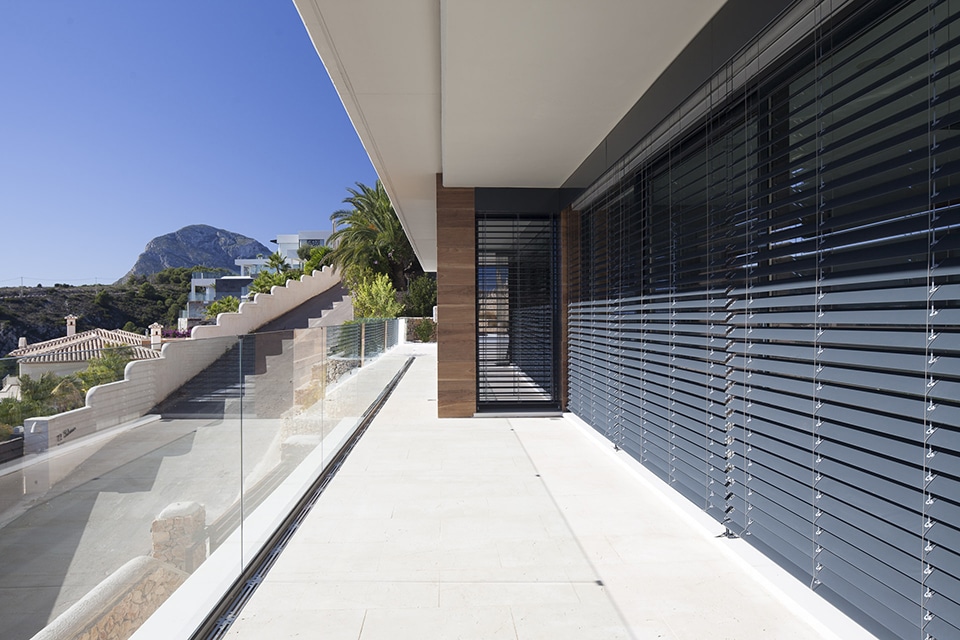 Terrasse moderne avec stores vénitiens gris, garde-corps en verre et vue panoramique sur une montagne.