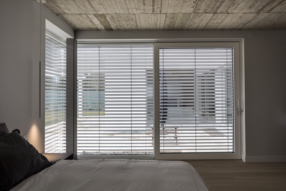 Chambre moderne avec plafond en béton brut et baies vitrées équipées de brise-soleil orientables Solomatic.