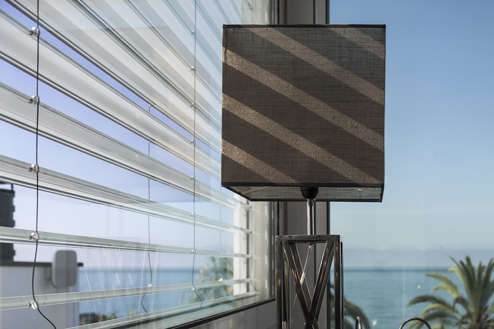 Lampe design et stores projetant des ombres, avec vue panoramique sur l'océan bleu et des palmiers tropicaux.