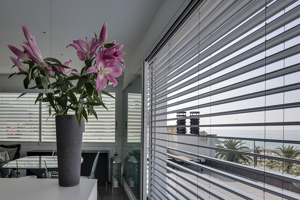 Lys roses en vase devant une baie vitrée avec stores et vue sur mer. Ambiance moderne et lumineuse.