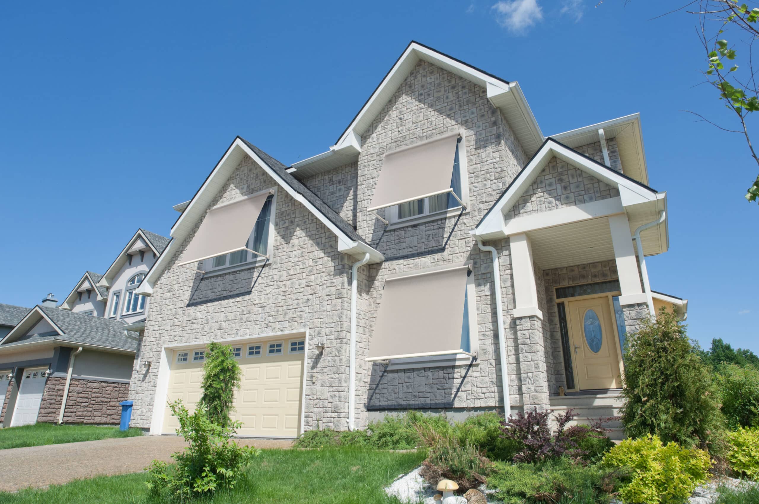 Maison moderne en pierre avec stores bannes extérieurs beiges, porte jaune et garage sous un ciel bleu.
