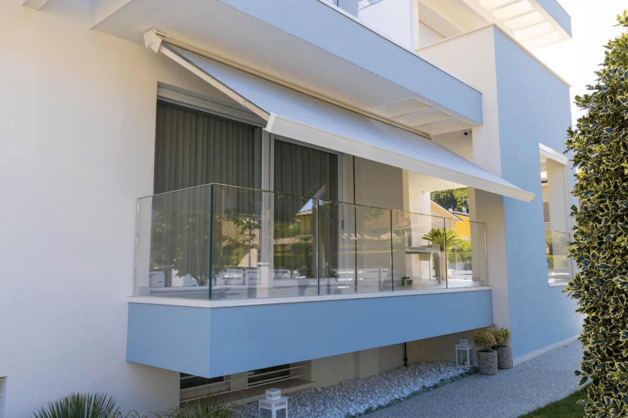 Store banne gris déployé sur un balcon moderne avec garde-corps en verre et façade bleu clair d'une maison.