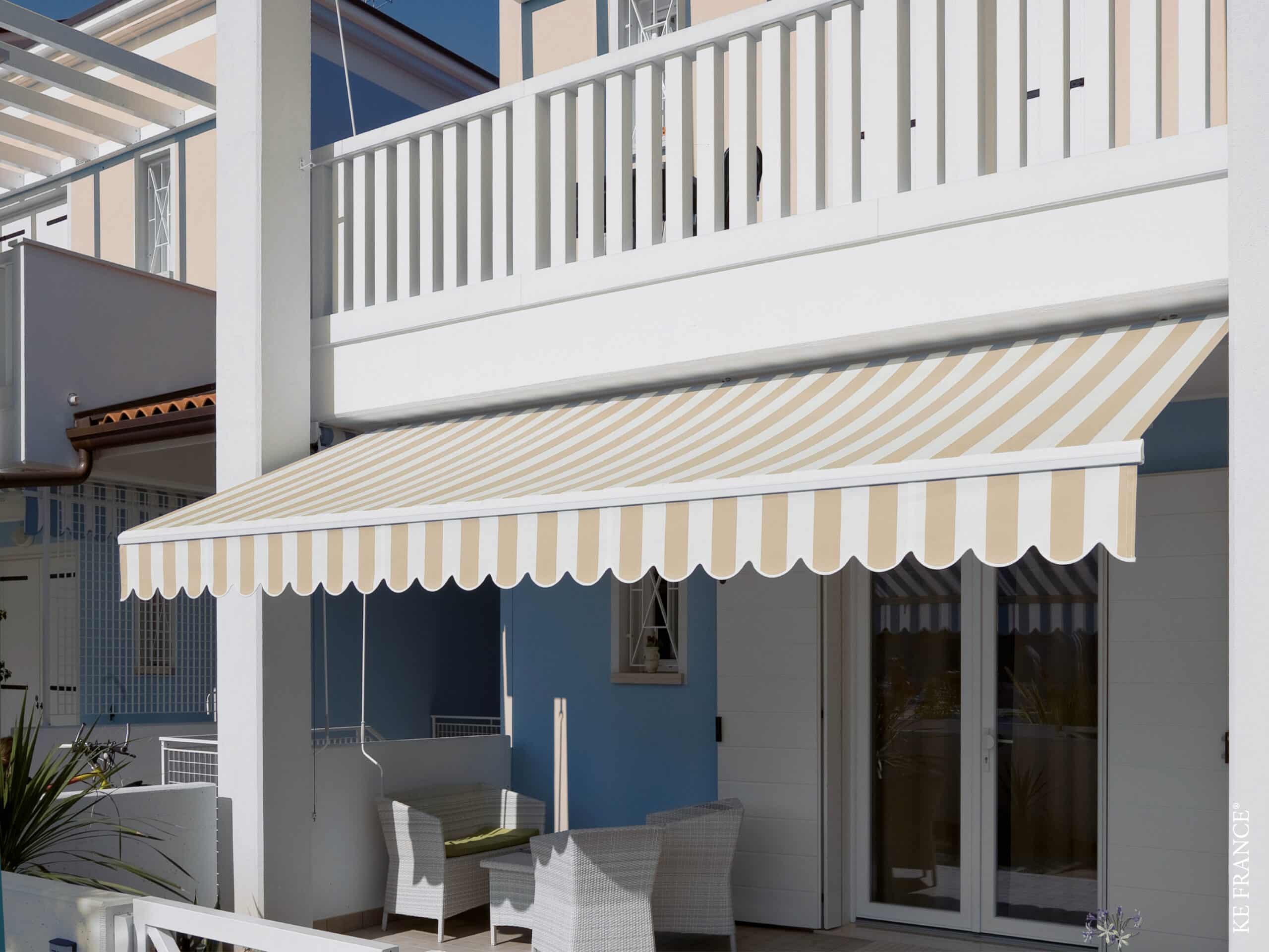 Store banne KE France rayé beige et blanc ombrageant un salon de jardin moderne sur une terrasse ensoleillée.