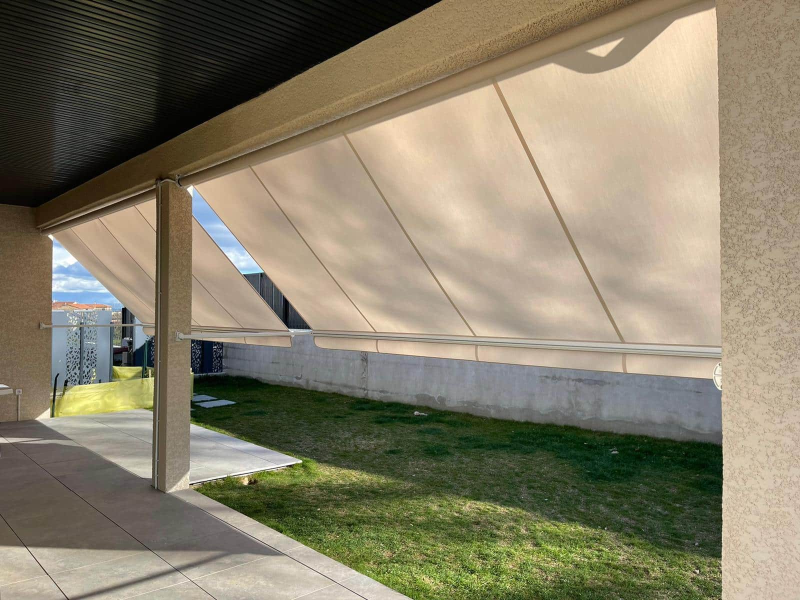 Stores bannes beiges déployés sur une terrasse carrelée devant un jardin avec pelouse et mur de clôture.