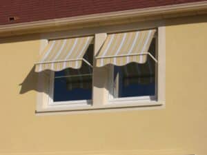 Deux fenêtres avec stores bannes rayés jaunes et gris sur une façade de maison ocre sous un toit de tuiles rouges.