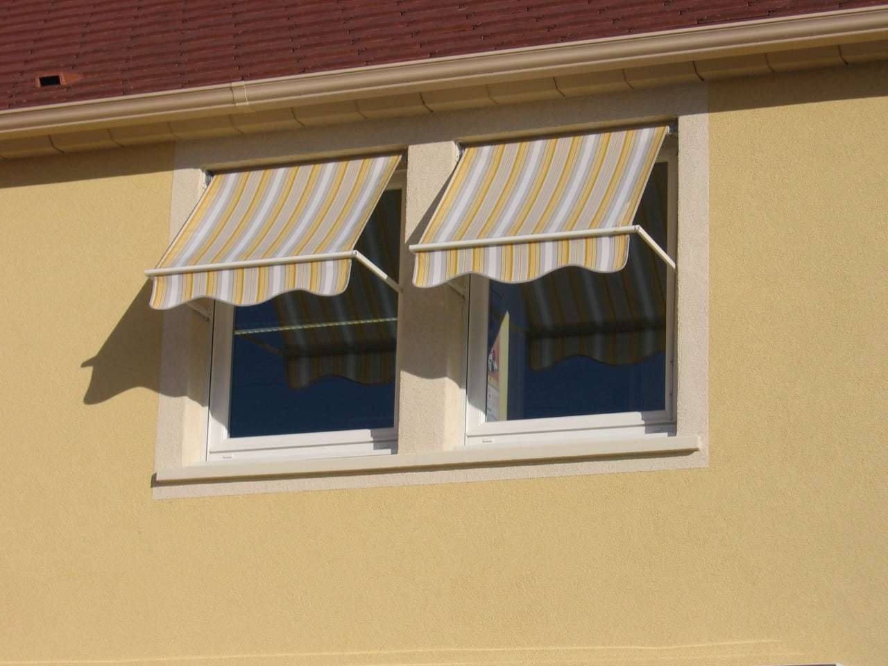 Deux fenêtres avec stores bannes rayés jaunes et gris sur une façade de maison ocre sous un toit de tuiles rouges.
