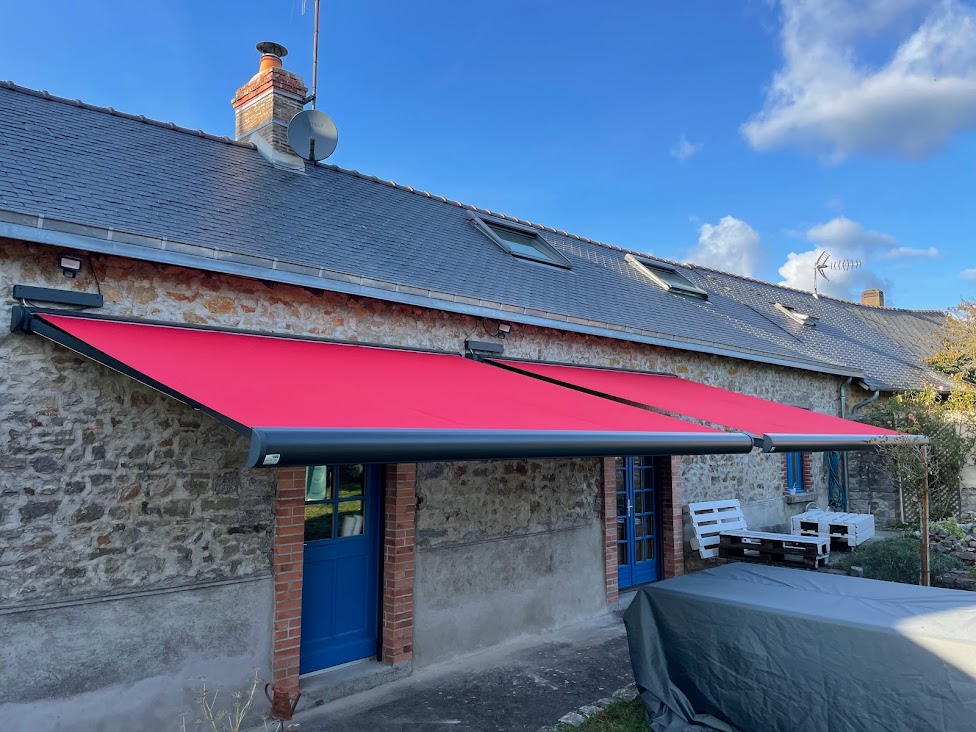 Store banne double rouge sur une maison en pierre avec portes bleues et toit en ardoise.