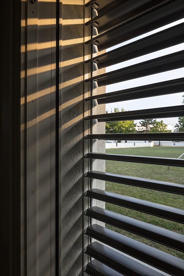 Store brise-soleil horizontal avec jeu d'ombres et lumière du soleil sur fenêtre bois, vue sur jardin.