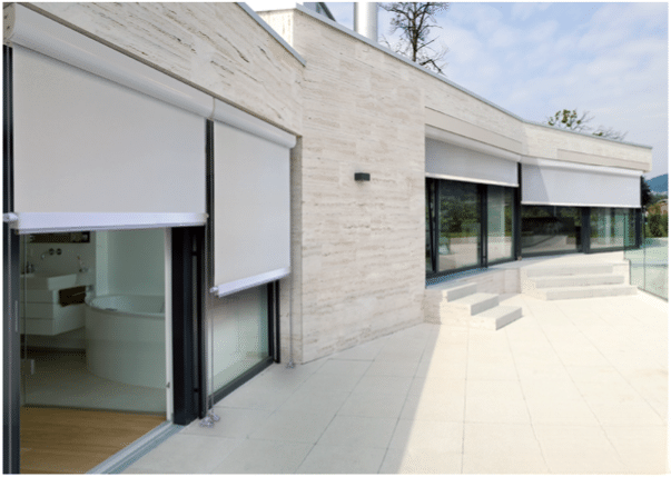 Stores enrouleurs extérieurs blancs sur terrasse moderne avec façade en pierre et grandes baies vitrées.