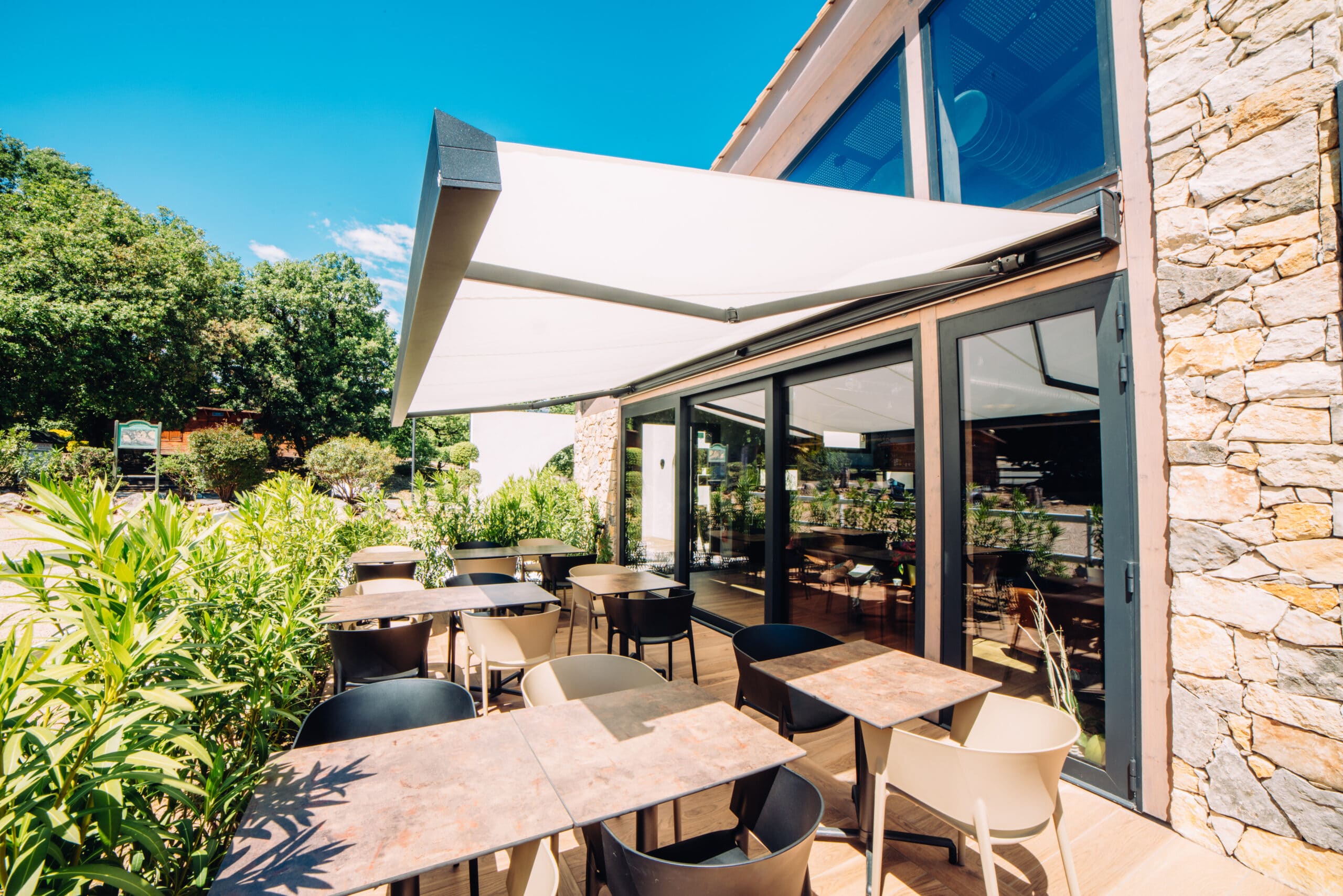 Terrasse moderne de restaurant avec tables, chaises et grand store banne blanc devant un mur en pierre naturelle.