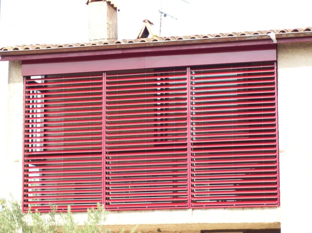 Brise-soleil orientables Grinotex rouges à trois sections installés sur une façade de maison sous un toit de tuiles.