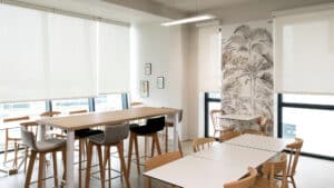 Salle de pause moderne avec tables hautes, chaises en bois et papier peint tropical dans un espace lumineux.