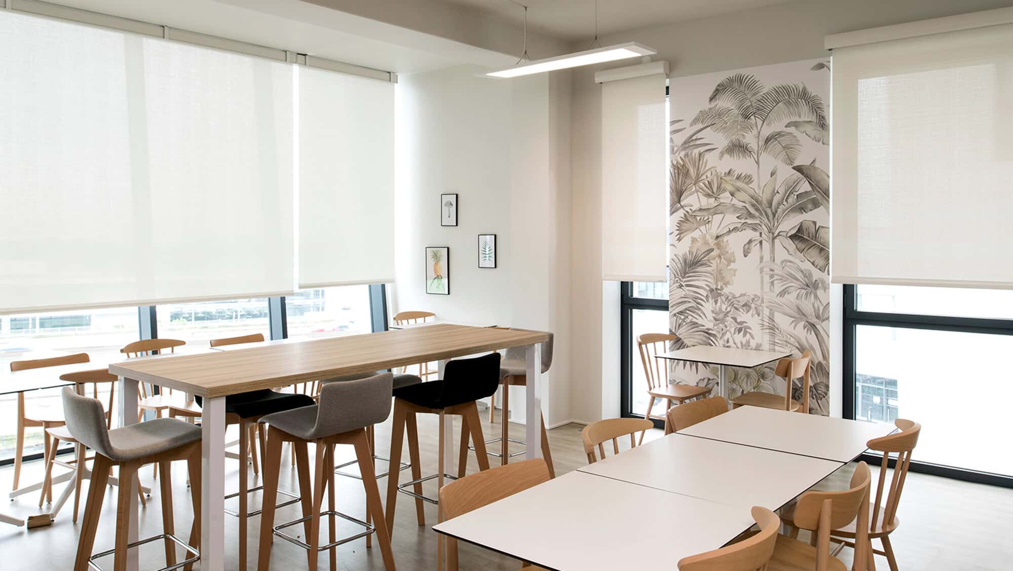 Salle de pause moderne avec tables hautes, chaises en bois et papier peint tropical dans un espace lumineux.