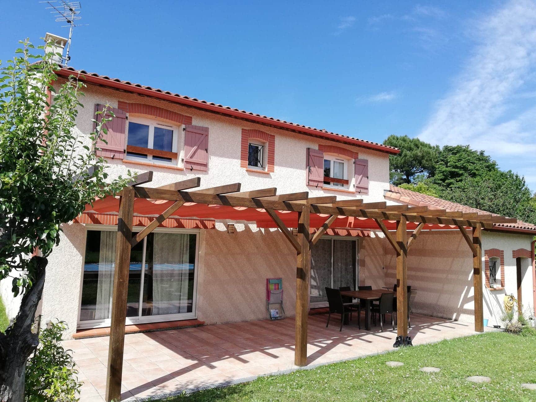 Maison avec terrasse carrelée, pergola en bois et toile Vélum et store orange sous un ciel bleu ensoleillé.