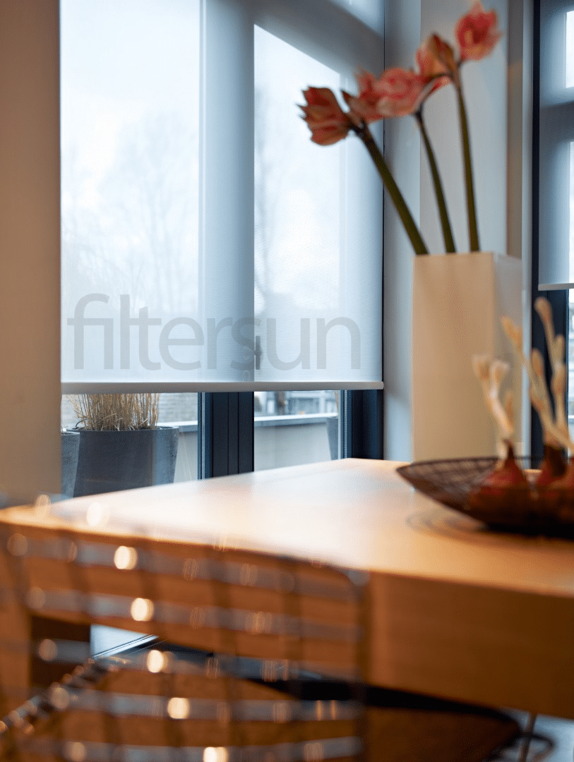 Store enrouleur tamisant Filtersun devant une fenêtre lumineuse, table en bois et vase de fleurs roses.
