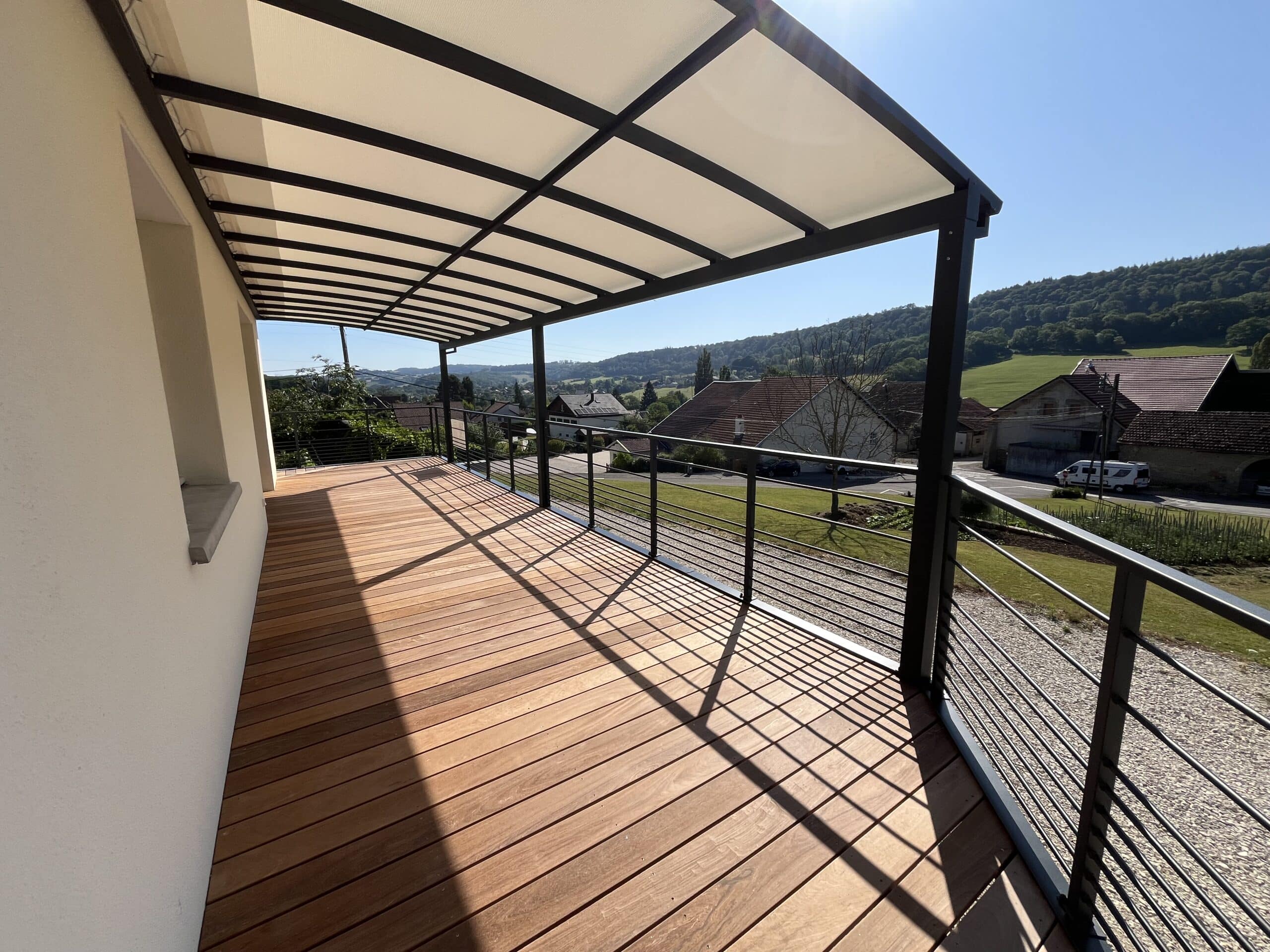 Terrasse en bois avec pergola noire et garde-corps métallique offrant une vue panoramique sur un village vallonné.