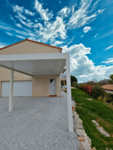 Carport en aluminium blanc devant une maison moderne, allée en gravier et jardin sous un ciel bleu nuageux.