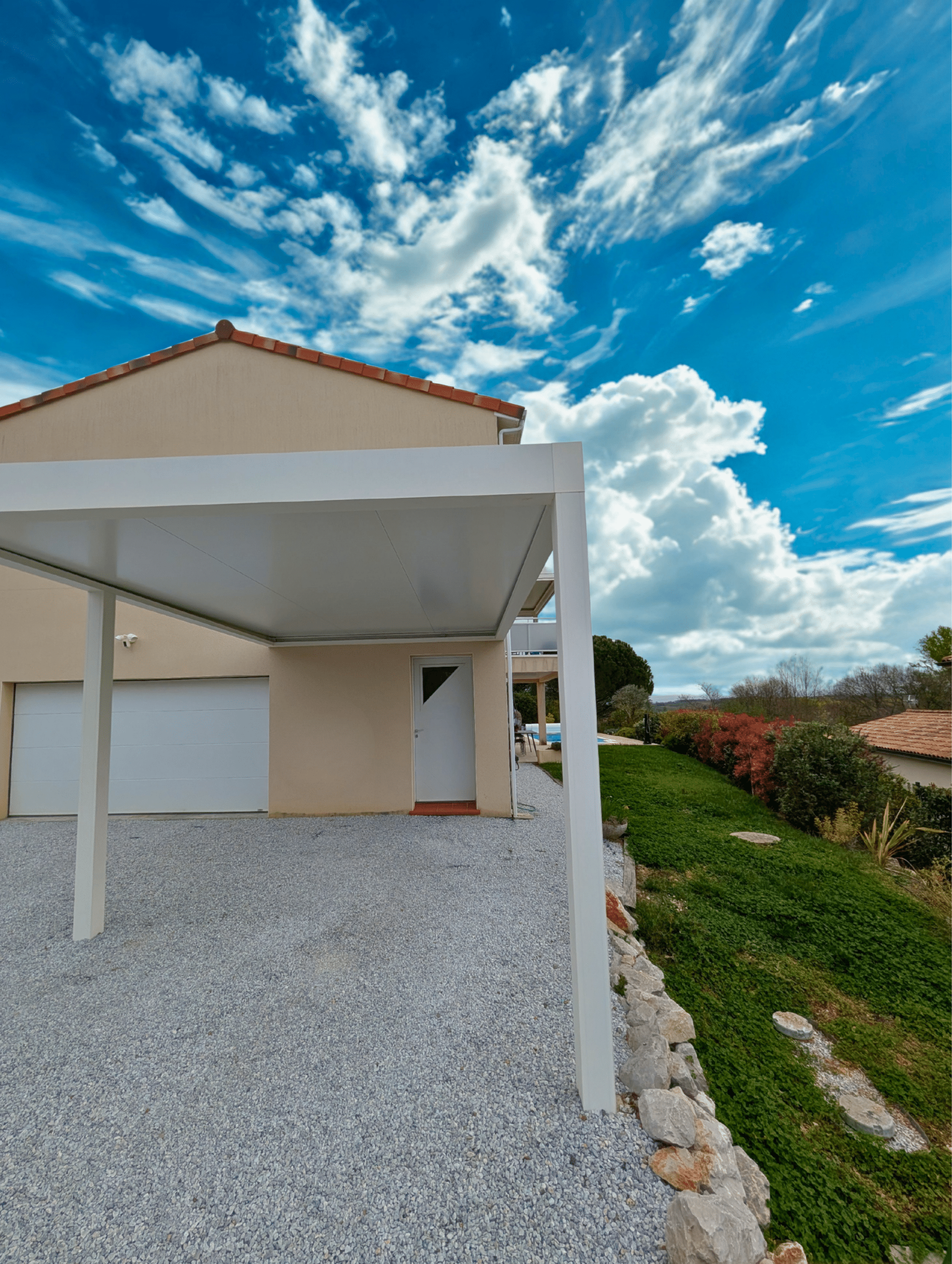 Carport en aluminium blanc devant une maison moderne, allée en gravier et jardin sous un ciel bleu nuageux.