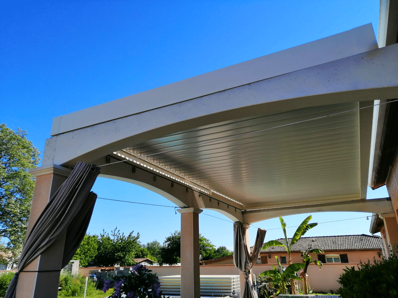 Pergola bioclimatique blanche à lames orientables avec rideaux gris sous ciel bleu.