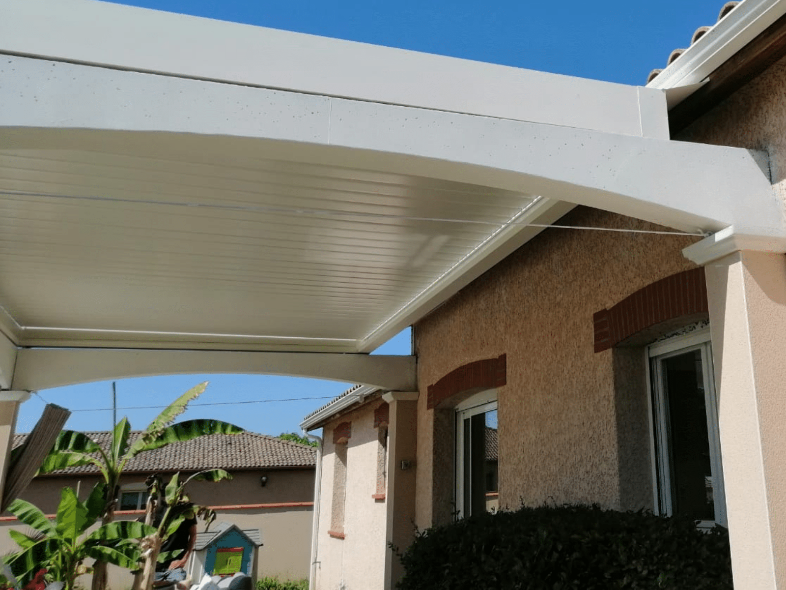 Pergola bioclimatique blanche adossée à une maison avec jardin tropical.