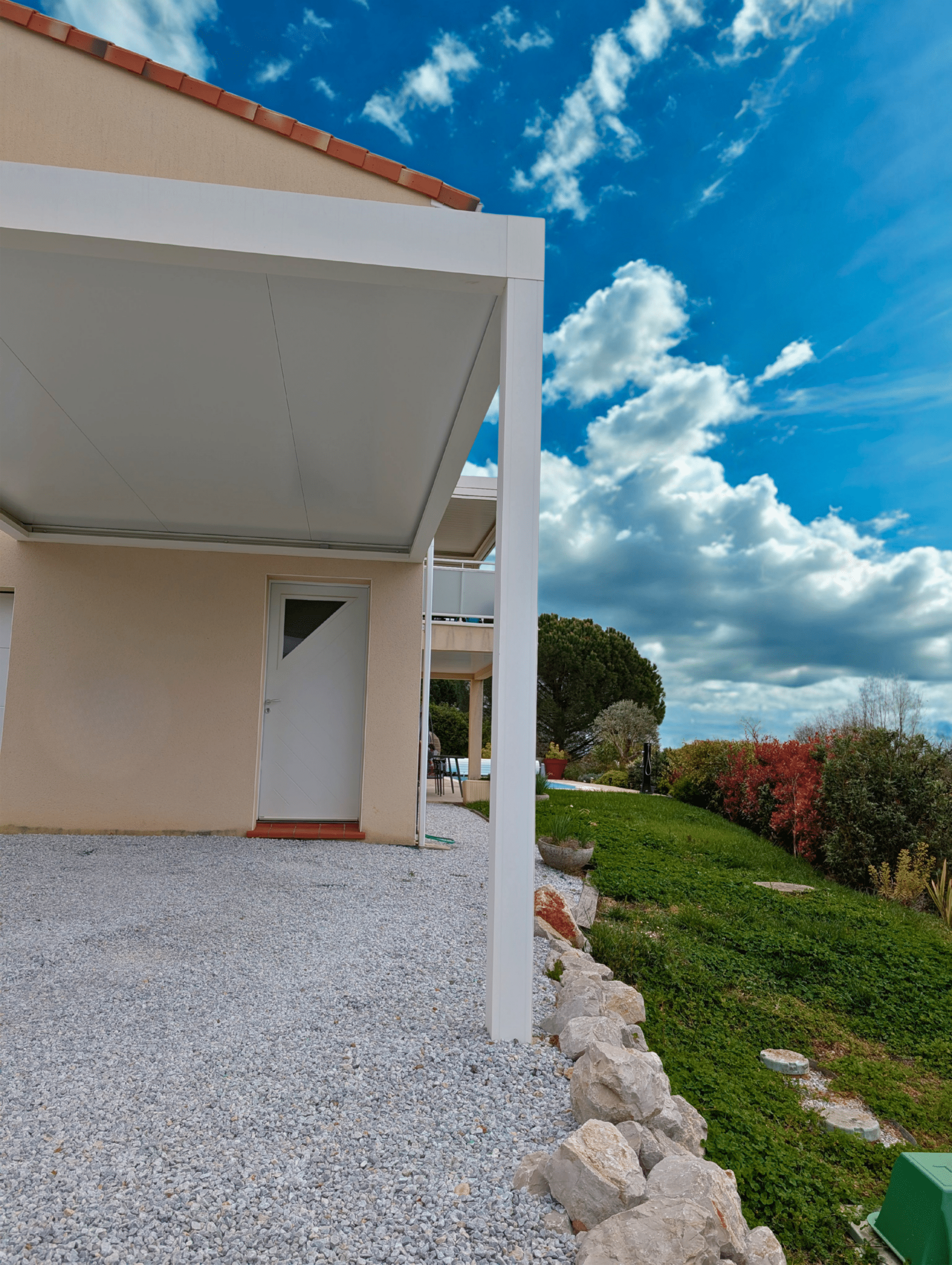 Pergola bioclimatique blanche adossée à une maison moderne, terrasse en gravier et jardin sous un ciel bleu éclatant.