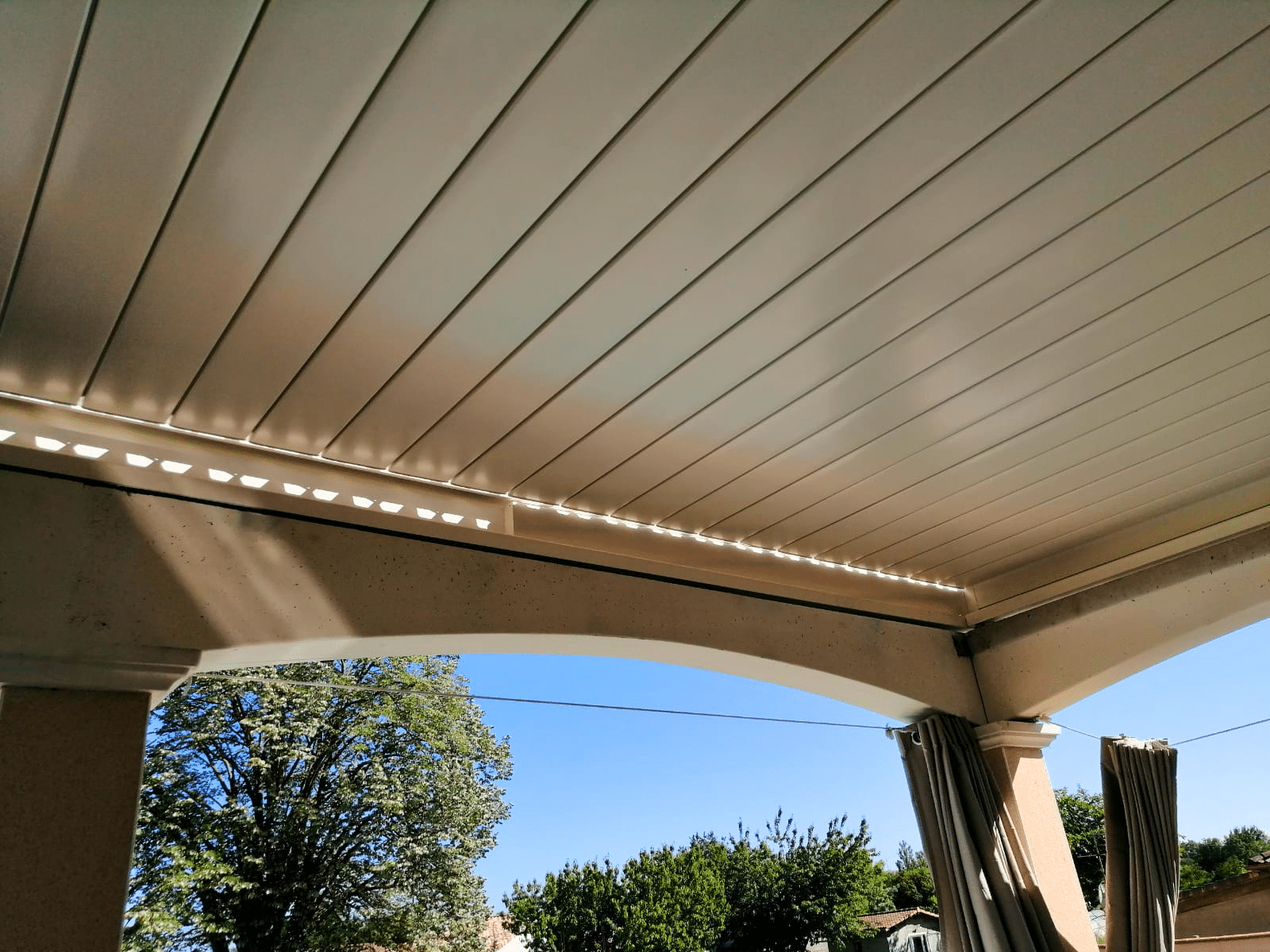 Pergola bioclimatique blanche fermée avec arches et rideaux extérieurs.